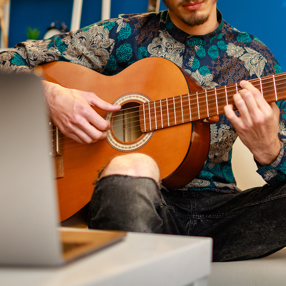 Joven caucásico elegante tomando clases de guitarra en línea en casa usando su computadora portátil